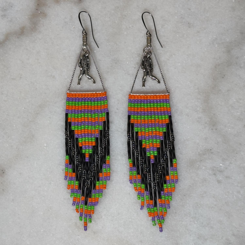 A pair of orange, purple, and green striped beaded fringe earrings, with zombie/monster charms on a marble background