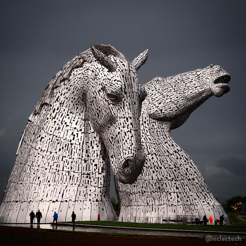 A square photo of The Kelpies, a steel sculpture, around 30 metres tall, of two horse heads. The one in front has its head down, the one behind is rearing up. They shine brightly against a dark grey sky. At the base you can see some people walking by, dwarfed by the sculptures.