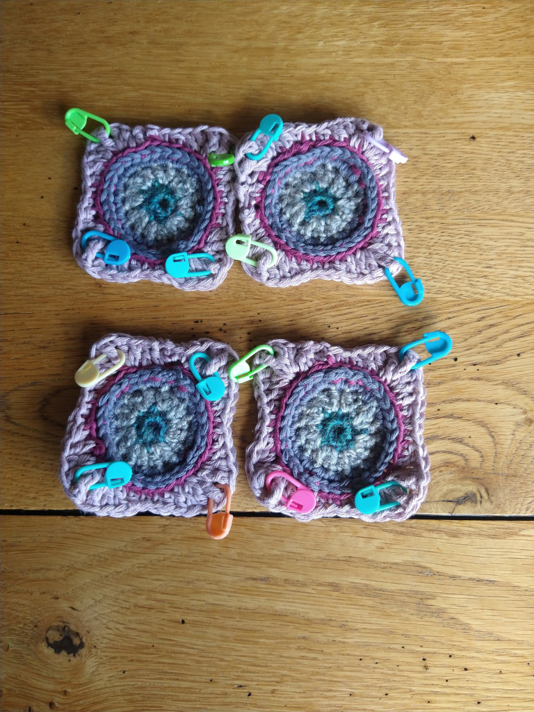 Four crochet squares in shades of pink and grey, with circle centres and coloured stitch markers at the corners