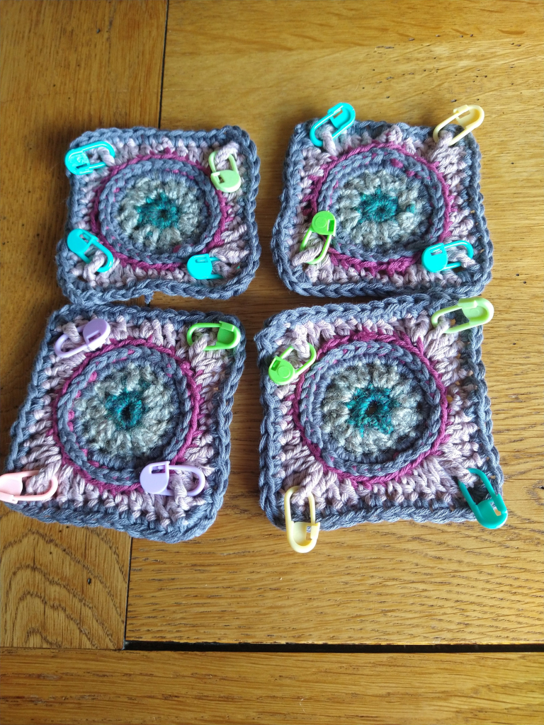 Four crochet squares in shades of pink and grey, with circle centres and coloured stitch markers at the corners