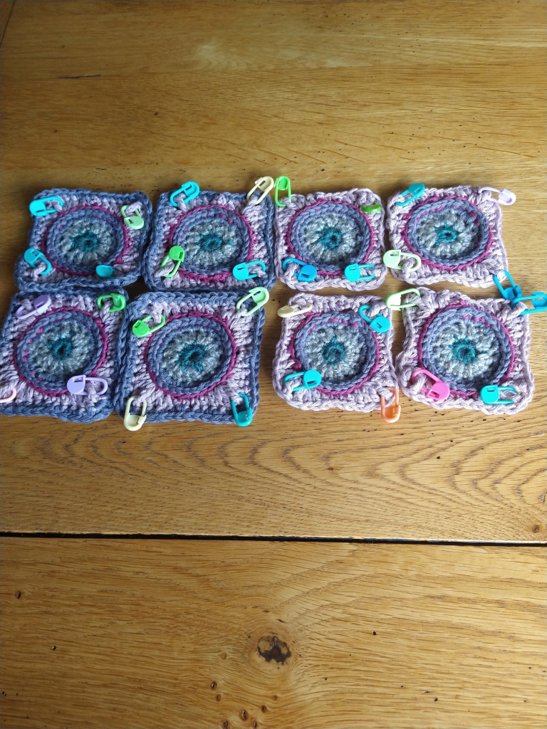 Eight crochet squares in shades of pink and grey, with circle centres and coloured stitch markers at the corners
Four are complete and four are waiting for their final round