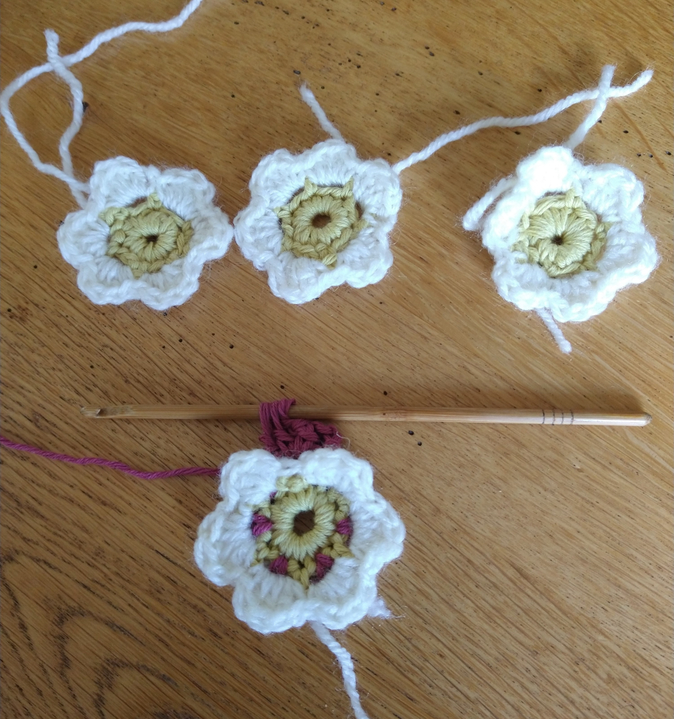 Four crochet flowers with yellow centres and white petals, at the bottom is a crochet hook attached to a flower currently being worked on in wine coloured yarn 