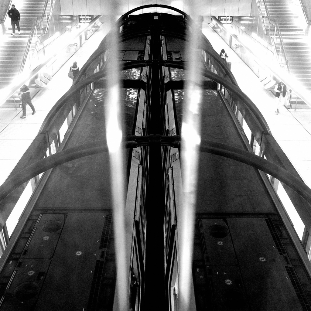 Photo de deux métros parisiens, prises d'une passerelle qui passe au-dessus de la voie. Les deux métros sont côte à côte, à l'arrêt. La passerelle est bordé de panneaux de verre séparés par un espace à travers lequel la photo a été prise. Les métros apparaissent plus nets dans l'interstice, tandis qu'ils sont un peu flous et pâles à travers les panneaux.