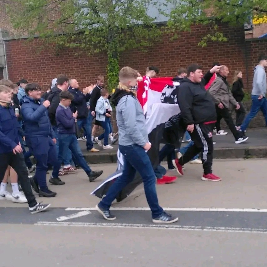 A crowd of mostly young, mostly male, Exeter City FC fans marching down a road, chanting and waving a banner saying something about Gary Caldwell 