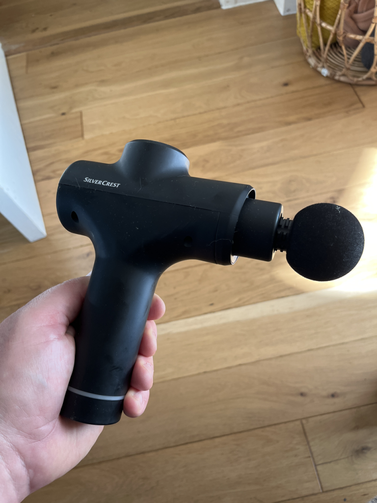 Photo of my hand holding a massage gun. In the background is laminate flooring. 