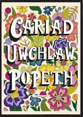 Multicoloured poster of leaves and flowers with the words CARIAD UWCHLAW POPETH (LOVE ABOVE ALL ELSE) in white capitals.