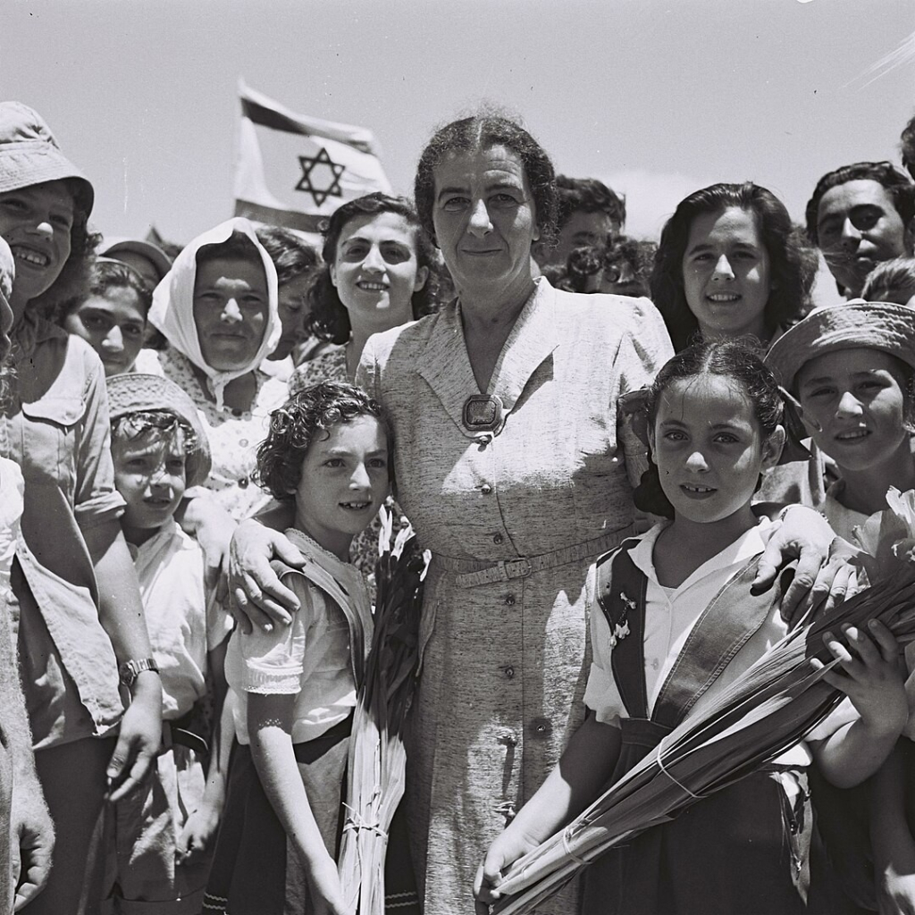 A photo of former Israeli Prime Minister, Golda Meir, with children on a kibbutz in 1950. She is standing with her arms around two girls with a crowd of mostly women behind her.

Photo credit: By Théodore Brauner - This is available from National Photo Collection of Israel, Photography dept. Government Press Office (link), under the digital ID D703-086.This tag does not indicate the copyright status of the attached work. A normal copyright tag is still required. See Commons:Licensing for more information., Public Domain, https://commons.wikimedia.org/w/index.php?curid=49704651