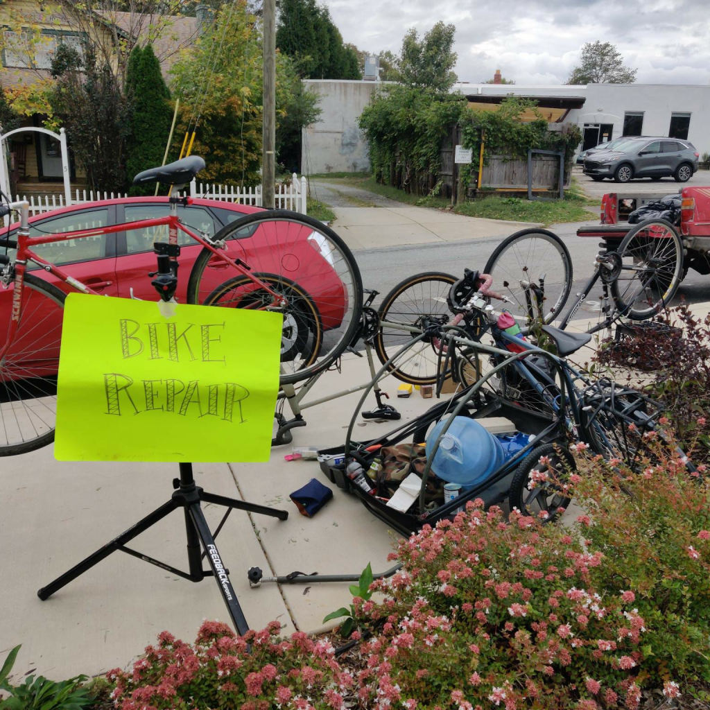 A photo of a bike repair clinic on the sidewalk outside Firestorm Books. Tools lie on the ground. Ared bike is suspended on a repair stand with an attached neon yellow sign that reads “BIKE REPAIR.”