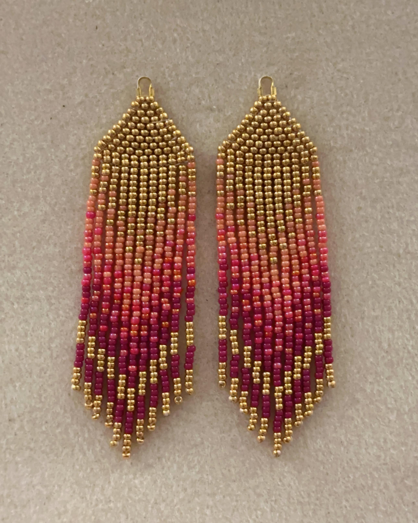 A pair of gold, peach, red, and purple ombre beaded fringe earrings on a bead mat