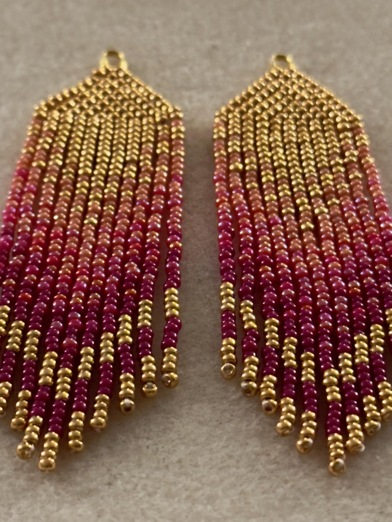 A close up of a pair of gold, peach, red, and purple ombre beaded fringe earrings on a bead mat