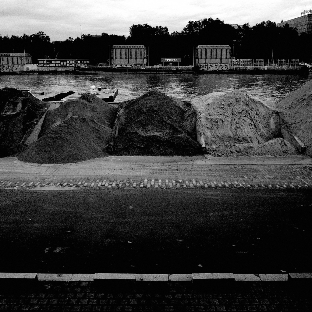 Photo de tas de sables de différentes couleurs, entreposés sur le bord de la Seine. En arrière plan, de l'autre côté du fleuve, des bâtiments qui ressemblent à des silos, devant une rangée d'arbres.