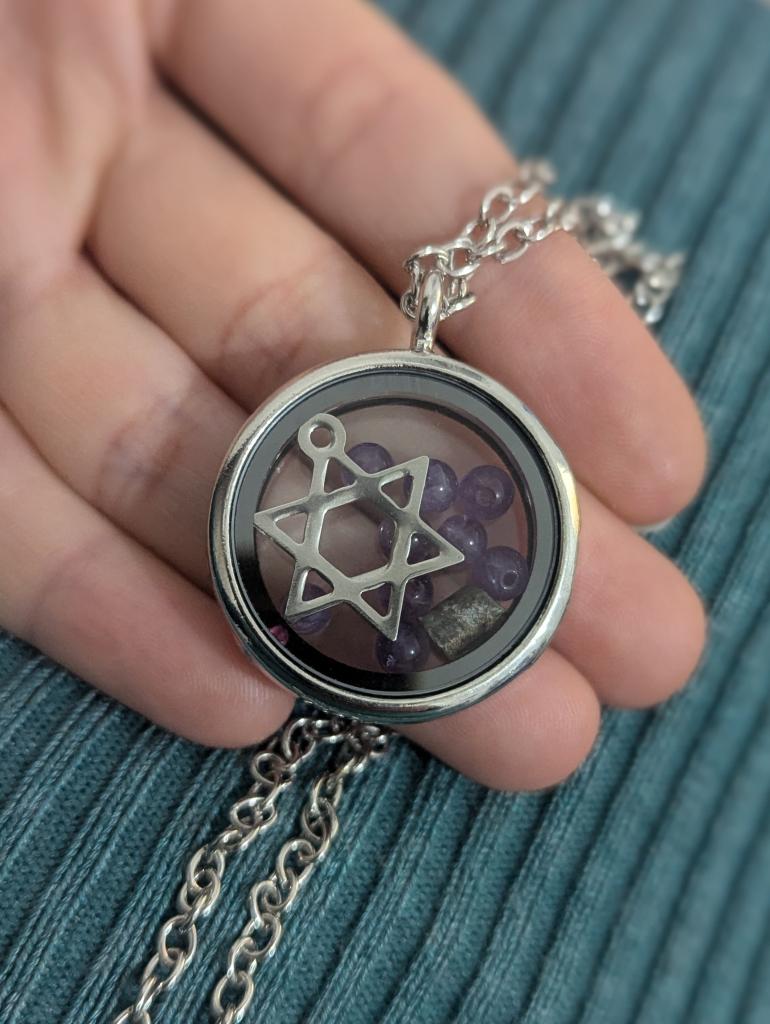 A photo of a round silver locked, in it a stainless steel Magen David, a piece of a grenade and many little round amethyst gemstones