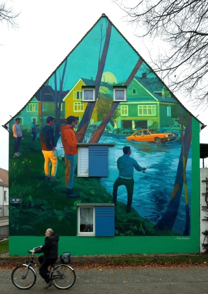 Streetartwall. A magnificent mural depicting a rushing river has been painted on the outside wall of a two-storey house. The image is positioned between four windows, painted in a naïve style and designed like an oil painting with bold colors and skillful brushstrokes. The mural shows a flood, a raging blue river on which an orange-colored car is being swept along and is now floating past the houses on the other bank.  Five dark-haired young men in modern, colorful clothing stand in the foreground on the bank between trees. With their hands in their pockets, they watch the scene impassively. Meanwhile, one of the men is fishing with a rod. It is unclear to the viewer whether the river has already burst its banks.
Info: The subject and motif were freely selectable. "The possible flooding is intended to reflect the uncertainty in society," explains the artist. Incidentally, the model for the five young men were his friends.