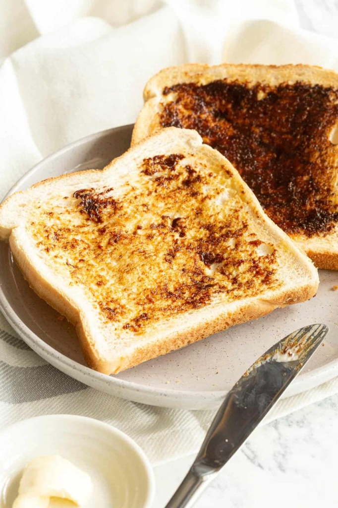 Two slices of toast on a plate. They have both been buttered and spread with Vegemite, one with a barely visible light spread, and one with a standard spread. A butter knife rests on the plate with margarine, Vegemite, and toast crumbs on the blade. 