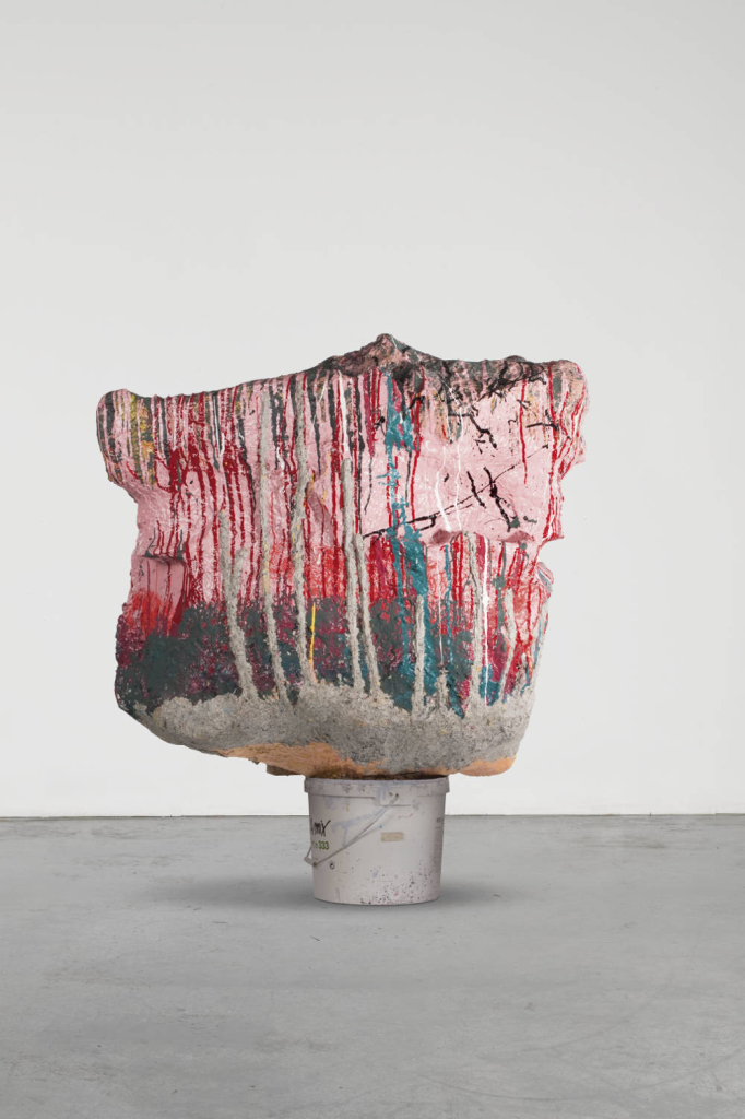 Abstract, textured sculpture with drippy pink, turquoise, and grey colors, balanced atop a white bucket