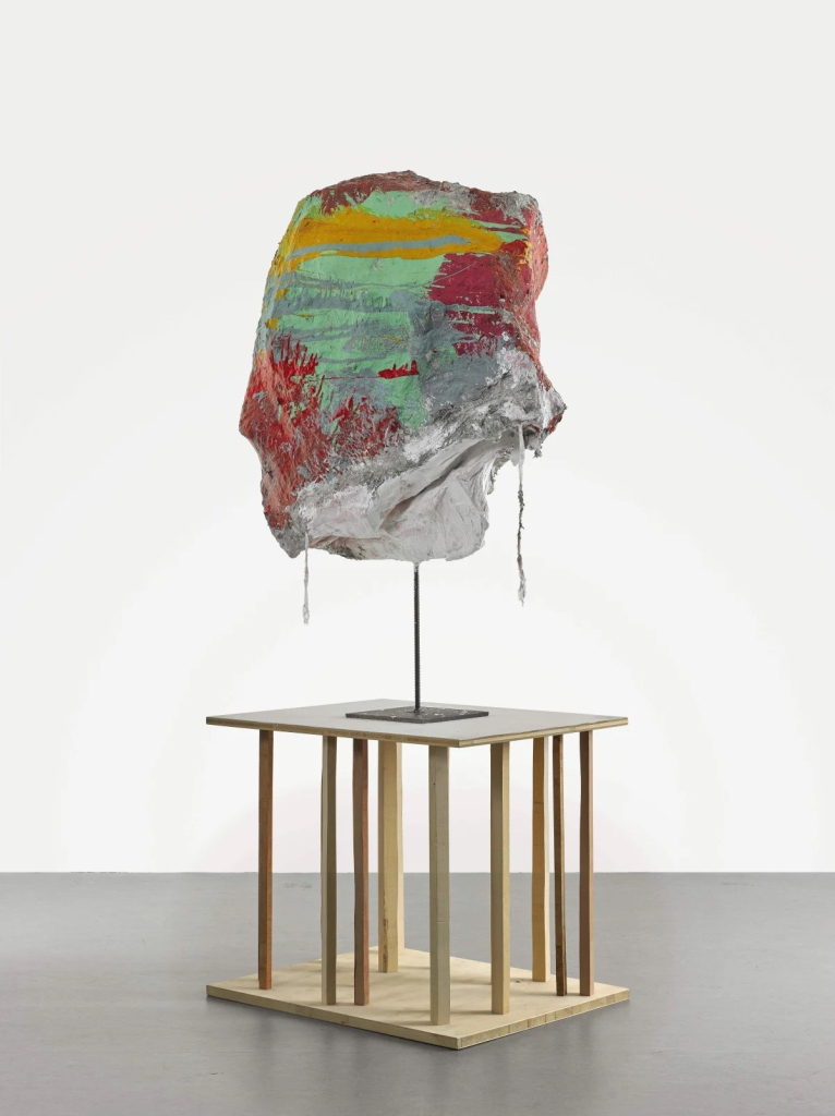 Abstract, textured sculpture with drippy grey, red, mint, and yellow colors atop a wooden plinth