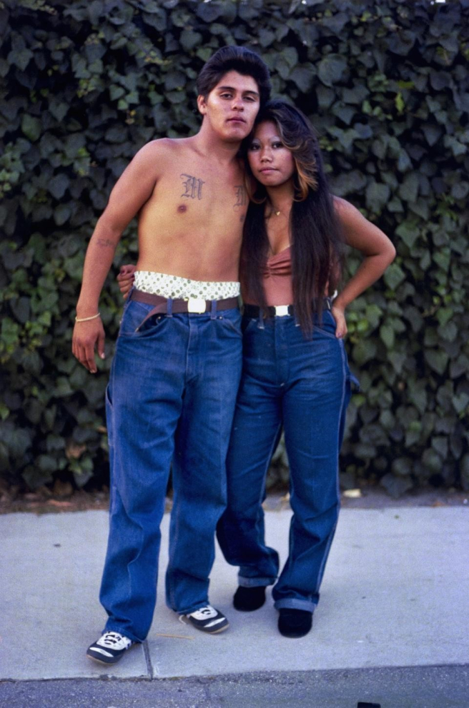 Portrait of a Latino man and woman standing with their arms around each other looking at the viewer, both wearing loose denim jeans, with a leafy wall behind them