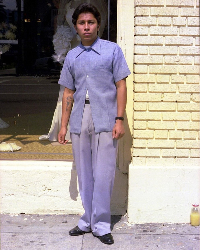 Photo of a young Latino man wearing a soft purple outfit and looking at the viewer, with a dress shop window behind him