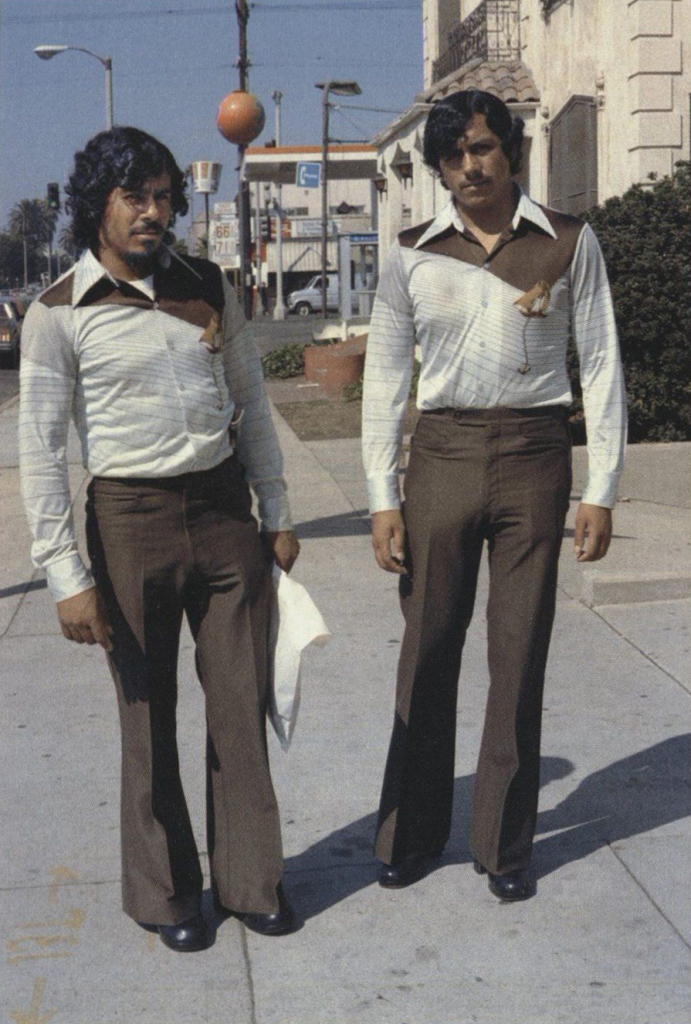 Photo of two Latino men standing on the street and wearing matching outfits - brown wide-leg pants and white and brown long sleeve button down shirts with large collars and naval insignias