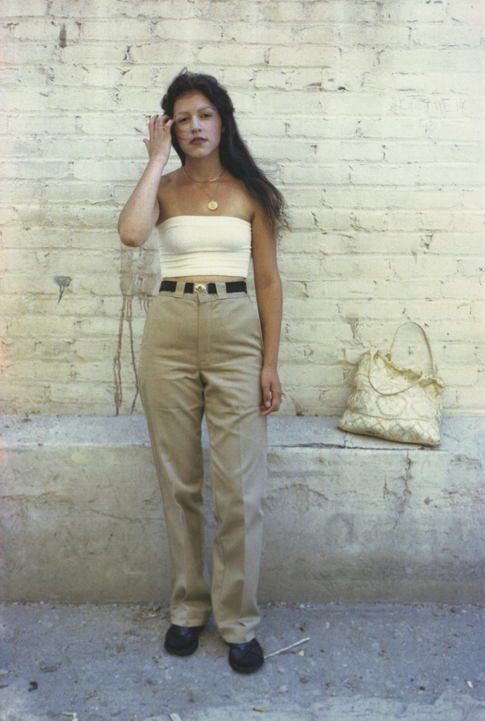 Photo of a young Latina woman standing against a sandy brick wall looking at the viewer, wearing waist-high khaki pants and a cream tube top, brushing her long hair out of her eyes with her right hand