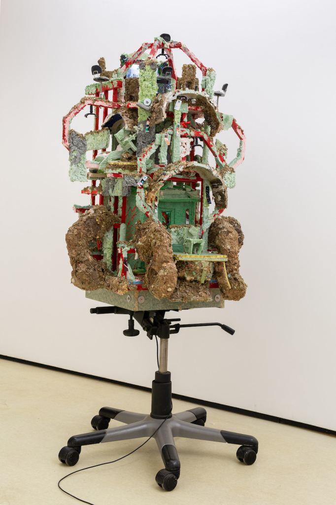 Mixed media sculpture of a textured green and brown structure sitting on top of a wheeled support like the bottom of an office chair