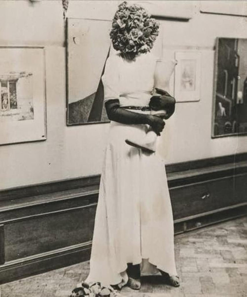 Black and white photo of a woman in a white gown and black gloves with her head obscured by a ball of roses, standing in an art gallery, holding a mannequin leg in her arms.