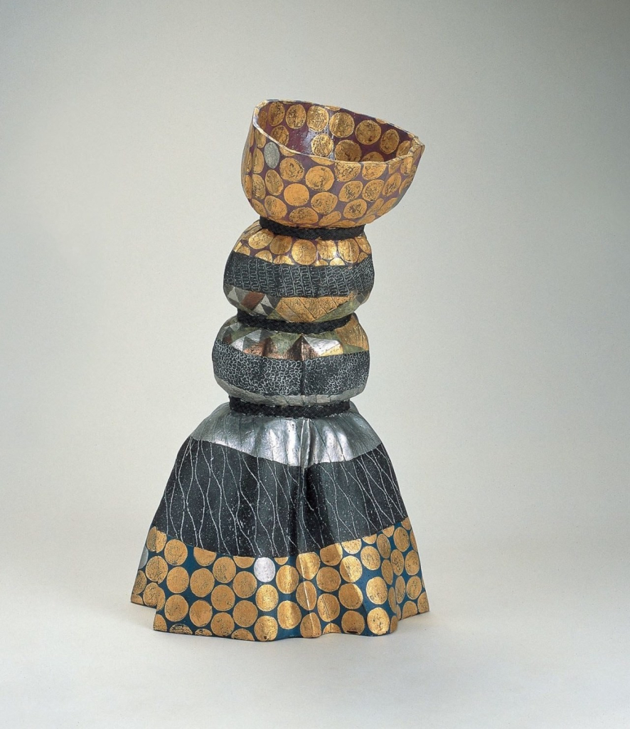 Ceramic sculpture resembling a standing skirt of draped, patterned fabric pulled in by three separate bands, with gold and silver embellishments and dark grey sections