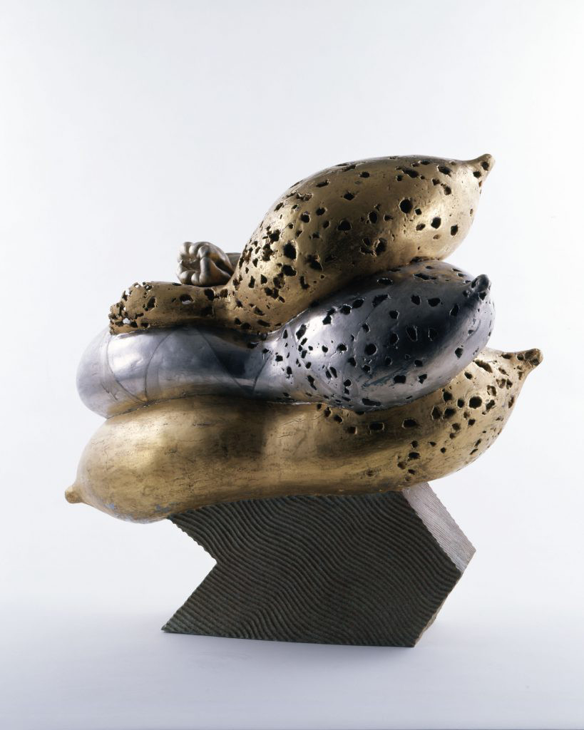 Ceramic sculpture of three abstract bulbous metallic forms with pockmarked holes lying atop a grooved stone base