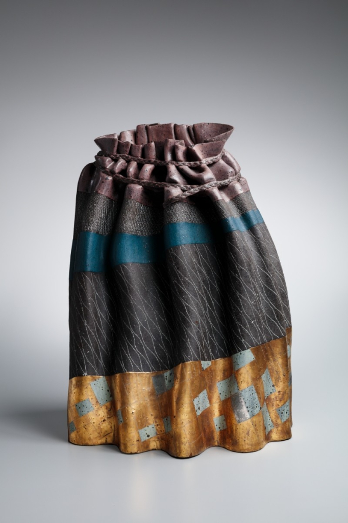 Ceramic sculpture resembling a standing skirt of loosely pleated fabric, with gold embellishments along the bottom and ruched upper section