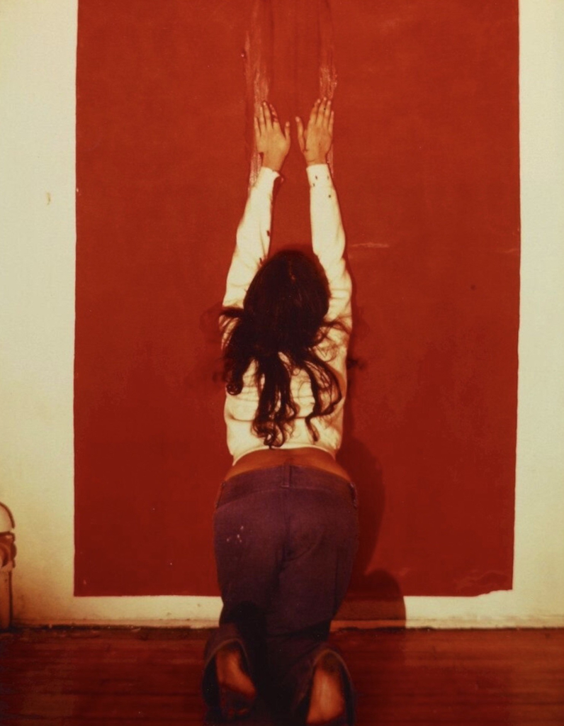 Photo of Mendieta kneeling in front of a canvas with her hands up, making gestural marks with her hands through a block of red pigment