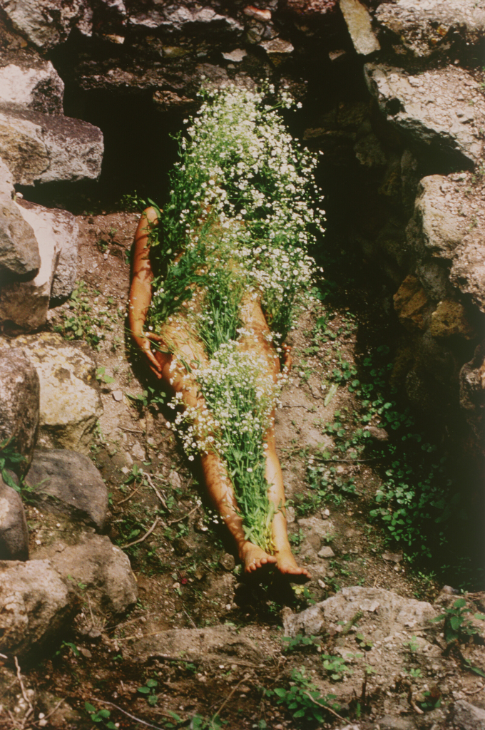 Photo of Mendieta posing lying down in a rocky hole, her nude body covered in green and white foliage