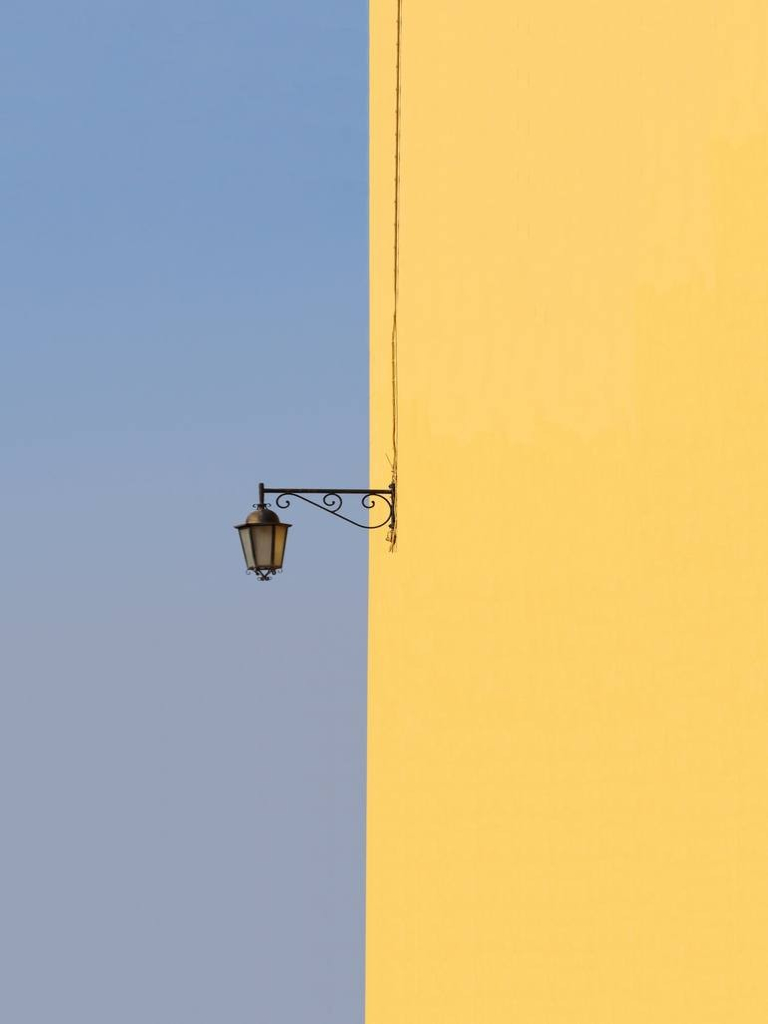 Photo of a yellow building on the right with a single wrought-iron lamp hanging off the wall, with clear blue sky on the left