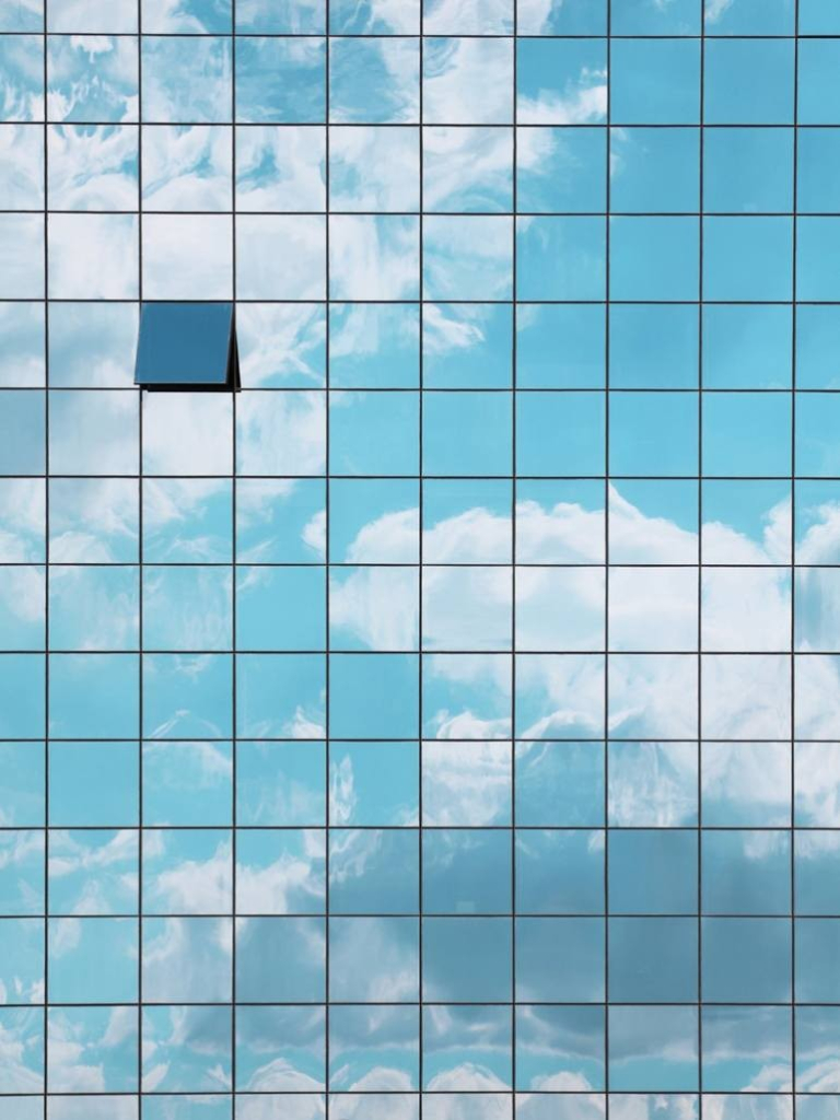 Photo of a gridded glass skyscraper wall reflecting clouds and blue sky, with one square window propped open