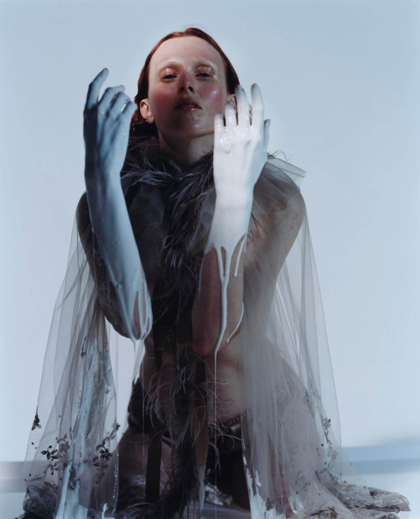 Photo of a red-haired white woman kneeling against a white sky wearing a gauzy loose cape, with hands held up in front of her body dripping white paint