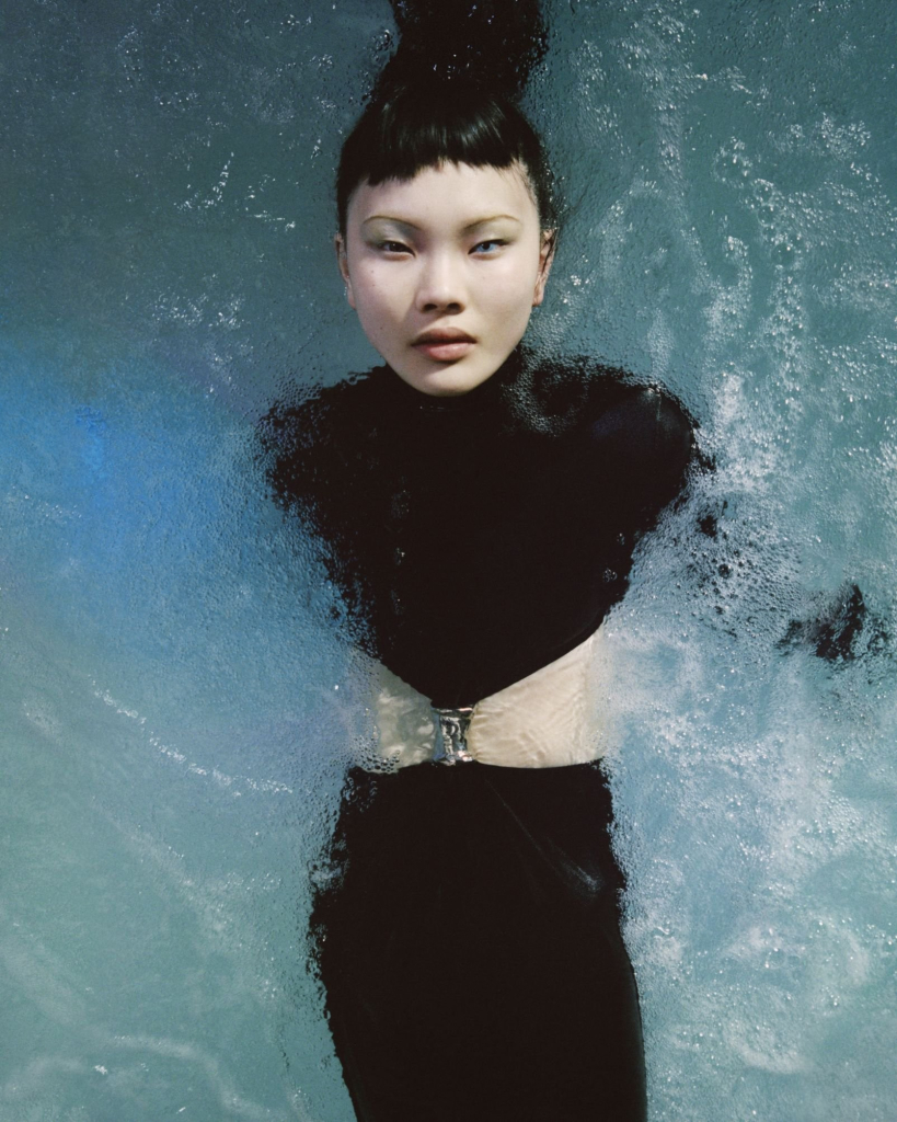 Photo of a young Asian woman partly submerged in foamy water, wearing a black two-piece dress and gazing directly at the viewer