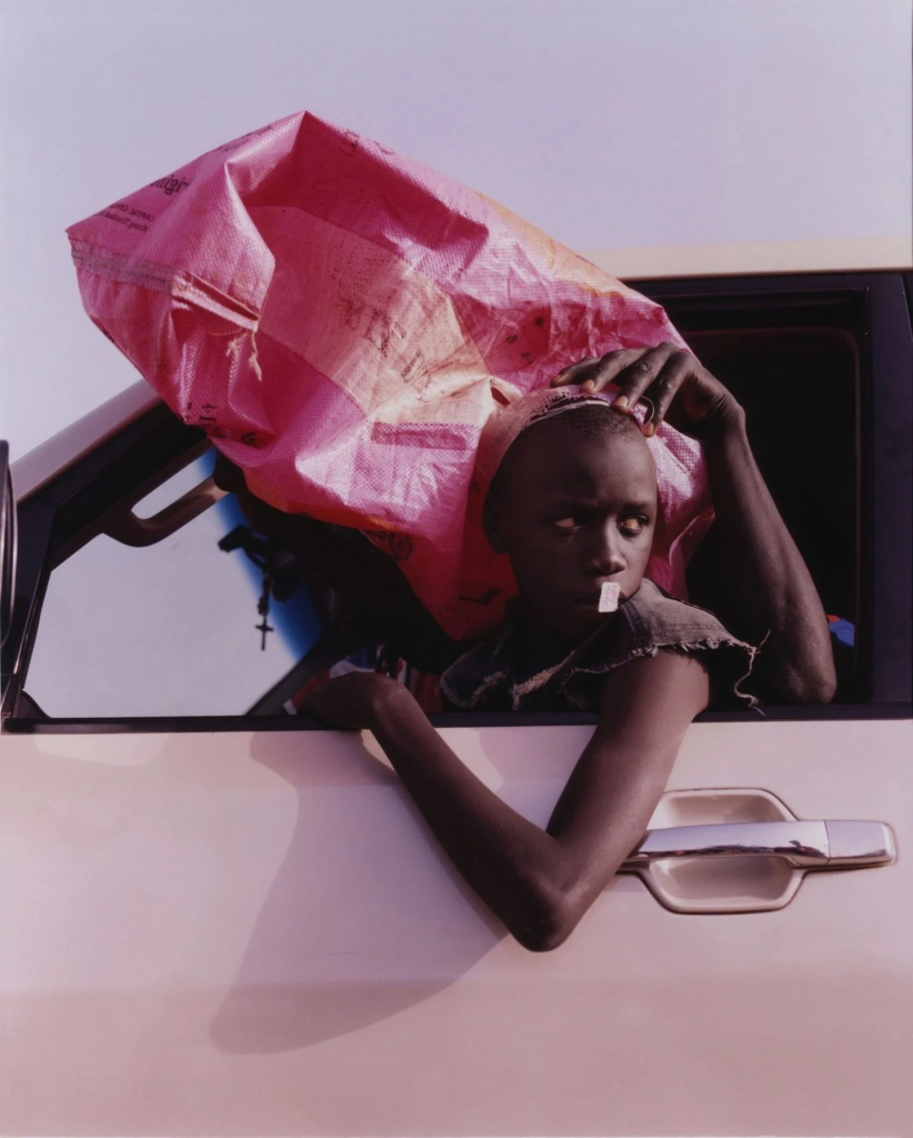 Photo of a young Black figure leaning out of a pink car, with a large pink bag pulled over their head by an unseen figure sitting in the passenger seat