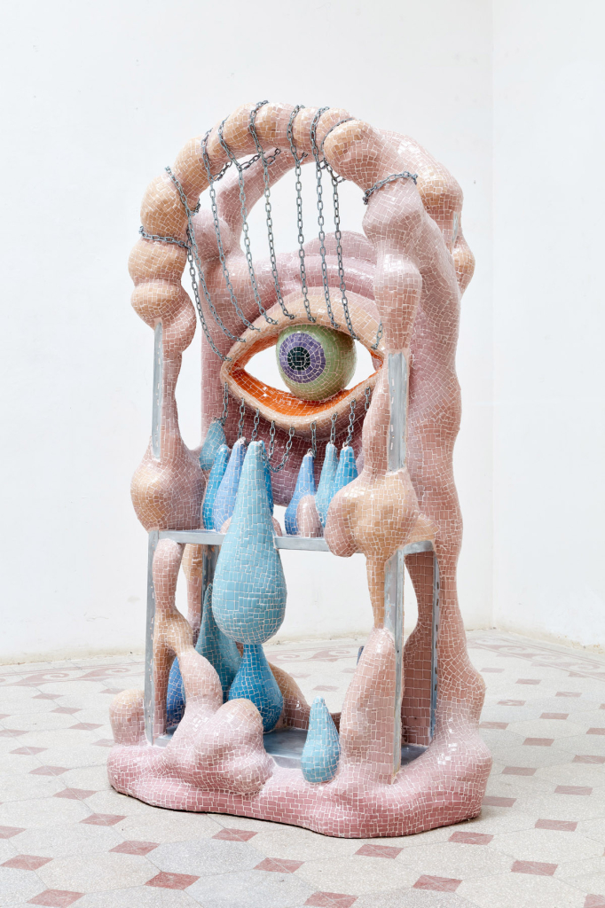 Abstract sculpture decorated with pink and blue mosaic tile, made of biomorphic forms with an array of chains and teardrop shapes, and a large green eye in the center