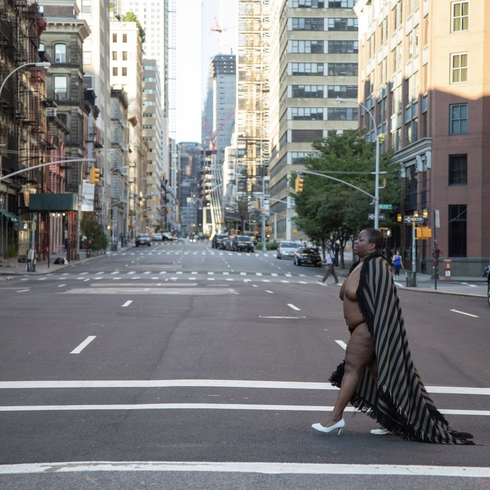 Photo of a Black woman in white heeled shoes nude underneath a long striped blanket, walking confidently across an empty street in NYC