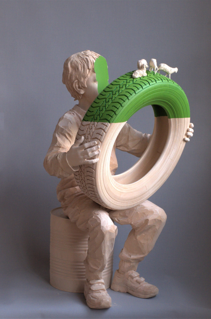 Wooden sculpture of a boy holding a car tire with small farm animals atop it, with a section of green paint bisecting the upper section of the tire and the front of the boy's face.