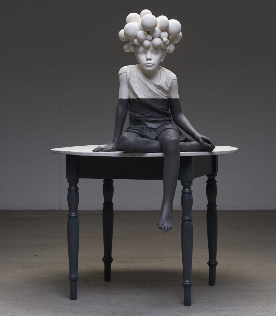 Carved wooden sculpture of a youth sitting on an ornate table. Their hair is replaced by differently sized balls as a large headdress. Their upper half is painted white, and below their elbows and chest painted black.