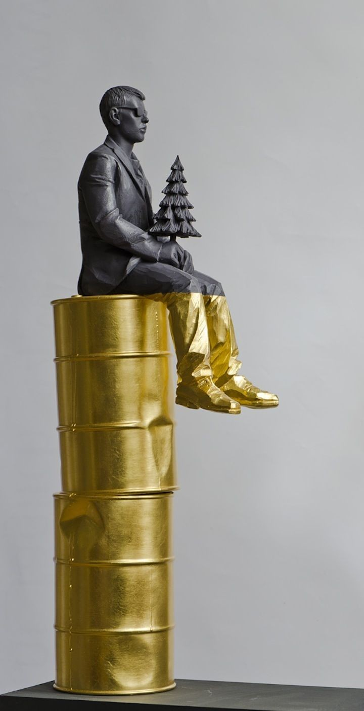 Sculpture of a man in a suit holding a small pine tree, all painted black, sitting on top of two stacked oil barrels painted gold. His lower legs and feet are dipped in gold as well.