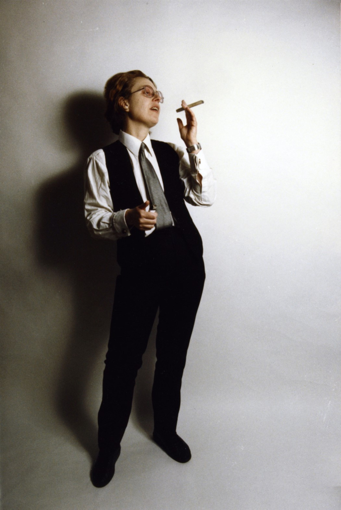 Full figure portrait of a white woman with short red hair and glasses, wearing black pants and vest over a white button down and tie, holding a cigar up to her face and leaning back in a confident pose against a white background