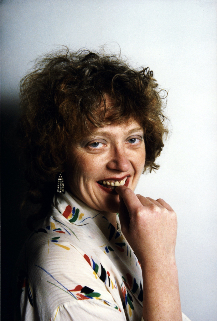 Portrait of a white woman with short red hair and glasses, wearing a floral blouse, posing with her hand up to her lower lip and smiling, against a white background