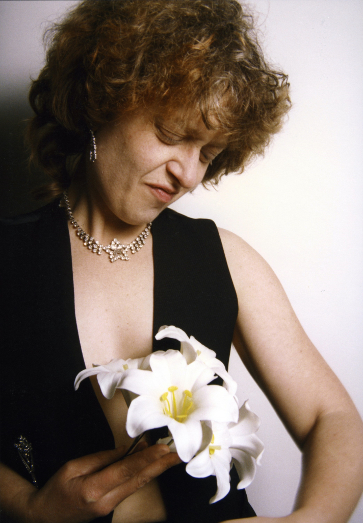 Portrait of a white woman with short red hair, wearing an open black vest and sparkly jewelry and holding white flowers up to her chest, looking down with a sneering expression, against a white background