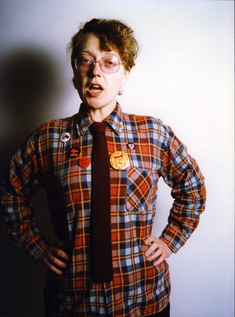 Portrait of a white woman with short red hair and glasses, wearing a red plaid shirt with pinback buttons and dark tie, posing with hands on hips and mouth open as if speaking, against a white background