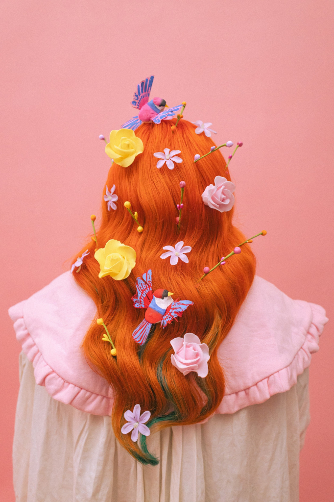 Photo of a figure facing away from the viewer, with long orange hair decorated with flowers and birds, against a peachy pink background