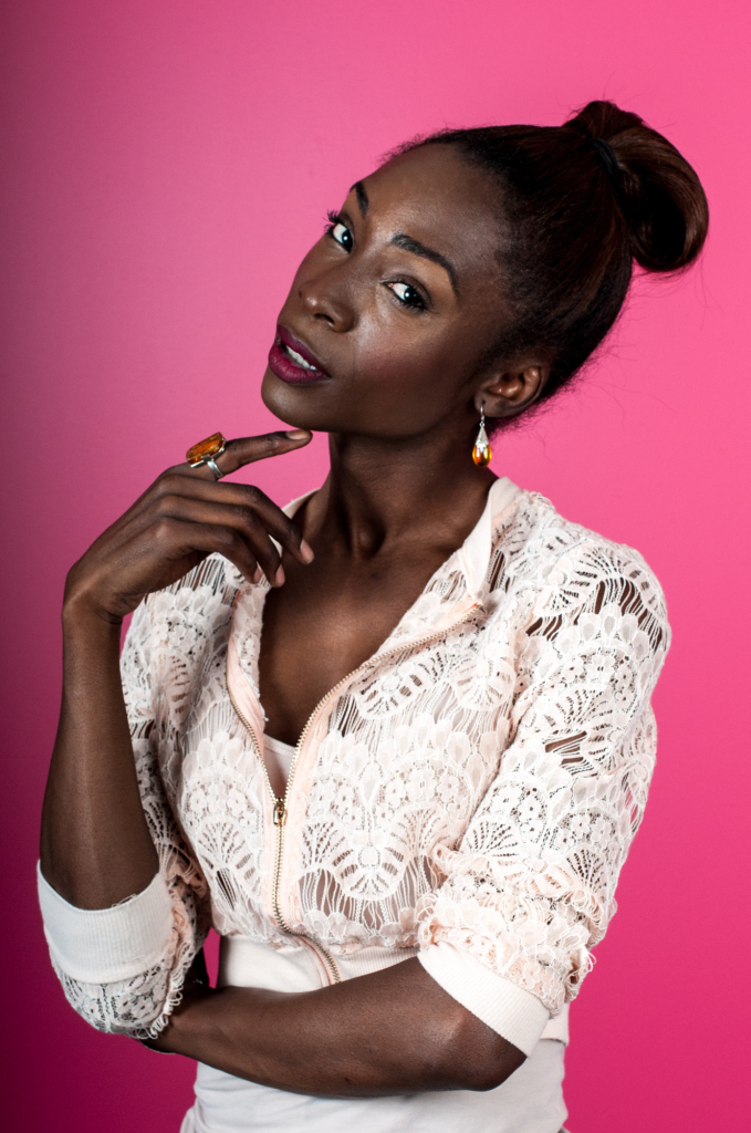 Photo of trans actress Angelica Ross posing with her hand up to her chin and her head tilted to the side, against a bright pink background