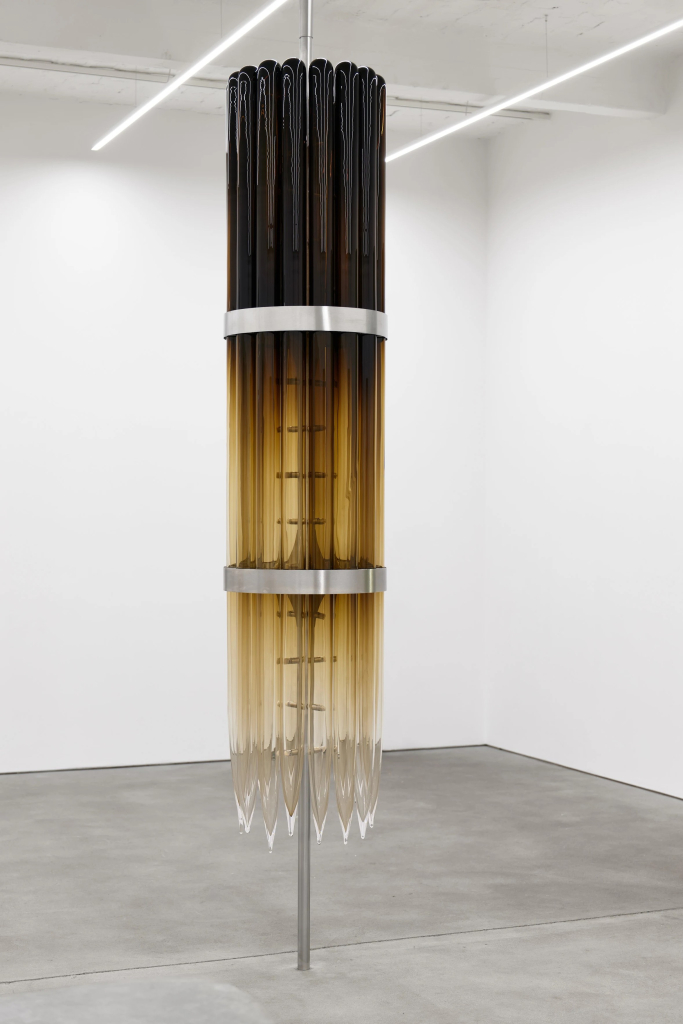 Hanging sculpture of a narrow cylindrical form made of long strips of glass with a dark brown-to-clear gradient