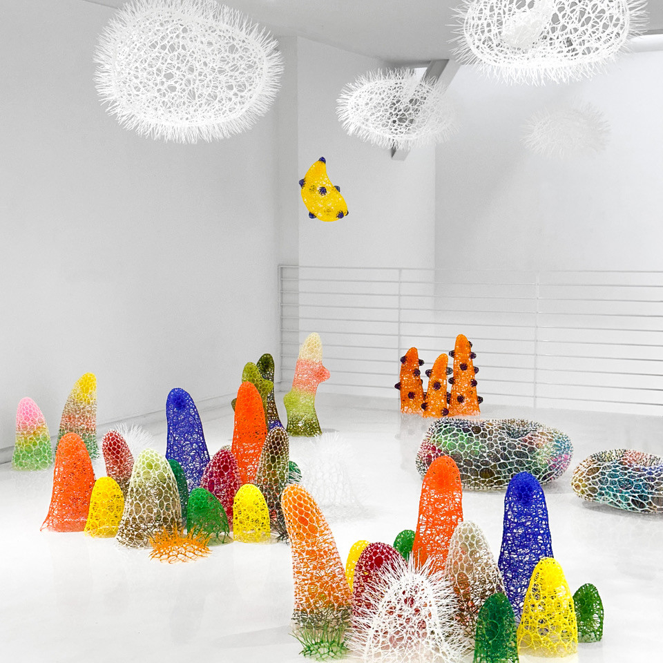 Installation of several freestanding rounded tower structures in different colors made of interlaced plastic, plus some white spiky orbs hanging from the ceiling, placed in a white gallery