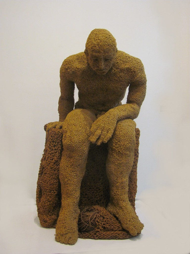 Light brown fiber sculpture of a seated man leaning forward with left on his lap